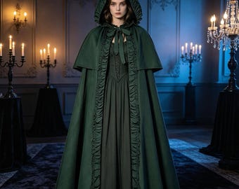 Può includere: Un mantello con cappuccio verde scuro che arriva fino a terra, caratterizzato da un bordo arricciato lungo l'apertura frontale e una chiusura a laccio sul collo. Il mantello è indossato sopra un abito verde scuro coordinato con corpetto.