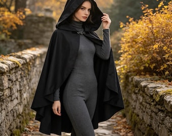Può includere: Un mantello nero con cappuccio dotato di una chiusura in metallo sullo scollo, indossato sopra un body a maniche lunghe color grigio antracite con collo alto. Il mantello presenta un cappuccio strutturato e un design aperto e fluido.