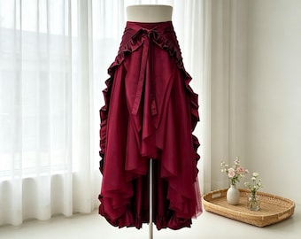Può includere: Una gonna asimmetrica color bordeaux con tessuto in tulle a strati, una vita strutturata con un laccio centrale e dettagli a balze lungo i pannelli frontali. Il capo è esposto su un manichino, mettendone in risalto la silhouette voluminosa.