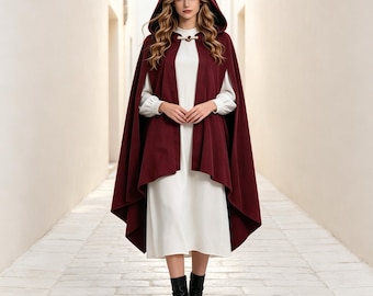 Può includere: Un mantello con cappuccio color bordeaux intenso con chiusura a levetta sul davanti, indossato sopra un abito bianco a maniche lunghe. Il mantello ricade sulle spalle fino a metà polpaccio, abbinato a stivaletti neri. Il capo è a tinta unita.