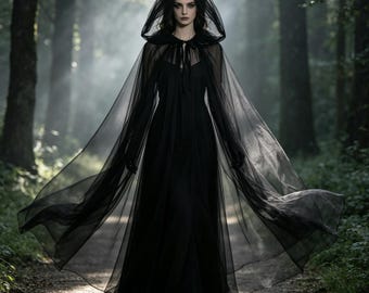 May include: A floor-length black dress featuring a sheer, flowing tulle cape with an attached hood. The cape is secured at the neck with thin black ties, creating a layered silhouette that drapes over the shoulders and extends outwards in a wide arc.
