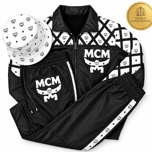 Op de afbeelding: Een zwart-wit trainingspak bestaande uit een jack met rits en een bijpassende broek met het MCM-logo en tekst. Het jack heeft een ruitpatroon op het bovenste gedeelte. Een witte bucket hat met een zwarte MCM-logoprint is inbegrepen.