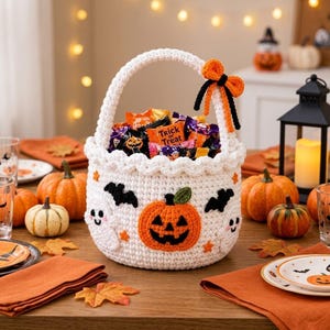 Puede incluir: Una cesta de ganchillo blanca con una calabaza naranja, dos fantasmas blancos y murciélagos negros. El asa está decorada con un lazo naranja y negro. La cesta está llena de varios envoltorios coloridos de dulces de Halloween.