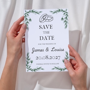 Op de afbeelding: Een witte Save the Date-kaart met een groene botanische rankrand, een lijntekening van twee in elkaar grijpende ringen en zwarte tekst: SAVE THE DATE FOR THE WEDDING OF James & Louise 20.08.2027 FORMAL INVITE TO FOLLOW.