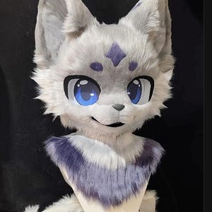 May include: A plush animal head mask featuring light gray faux fur, large blue eyes with black outlines, and a dark purple chest patch. The forehead displays a purple diamond-shaped marking with two smaller dots, and the ears are pointed.