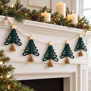 May include: A macrame garland featuring five dark green rope Christmas trees suspended from a thick white cord. Each tree is topped with a light-colored wooden star and finished with a small tan tassel at the base of the trunk.