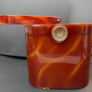 May include: A rectangular handbag with a glossy, marbled amber and brown finish. The bag features a hinged top handle and a circular, gold-toned metal clasp with a detailed, radiating sunburst or fan-like pattern centered on the front lid.