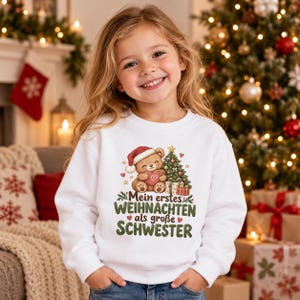 May include: A white long-sleeve sweatshirt featuring a graphic of a brown teddy bear wearing a Santa hat and holding a heart, next to a decorated Christmas tree and gifts, with the German text Mein erstes WEIHNACHTEN als große SCHWESTER.
