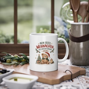 Puede incluir: Una taza de cerámica blanca con el gráfico de un oso de peluche con gorro de Papá Noel y bufanda verde, sosteniendo una estrella. El texto dice "MEIN ERSTES WEIHNACHTEN ALS GROSSER BRUDER 2026" con ilustraciones de árbol, regalos y farol.