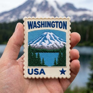 May include: A rectangular embroidered patch shaped like a postage stamp featuring the text WASHINGTON at the top and USA with a star at the bottom. The center depicts a snow-capped mountain, a blue lake, and dark green pine trees.