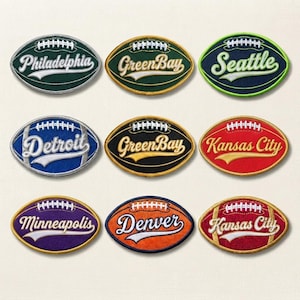 May include: Nine oval-shaped embroidered patches shaped like American footballs. Each patch features a city name in white or gold script, a white football lace graphic, and a colored border. The cities include Philadelphia, Green Bay, Seattle, Detroit, Kansas City, Minneapolis, and Denver.