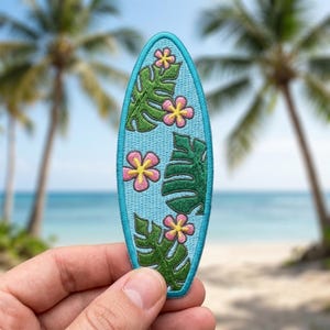 May include: An embroidered patch shaped like a surfboard with a light blue background. The design features green monstera leaves and pink flowers with yellow centers. The patch has a solid light blue stitched border around the entire edge.