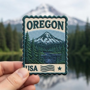 May include: A rectangular embroidered patch with a scalloped edge featuring a mountain landscape, a lake, and evergreen trees. The text OREGON is at the top in dark green, with USA, wavy lines, and a star at the bottom on a cream background.