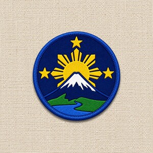 May include: A circular embroidered patch featuring a dark blue background with a yellow sun and three yellow stars. Below the sun is a white snow-capped mountain peak, green hills, and a blue river, all enclosed by a thick blue stitched border.