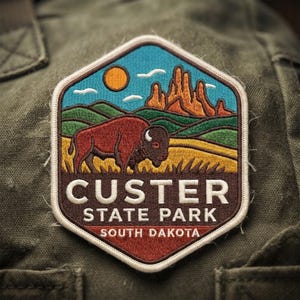 May include: A hexagonal embroidered patch featuring a brown bison standing in a yellow field with green hills, orange rock formations, and a yellow sun against a blue sky. The text reads CUSTER STATE PARK SOUTH DAKOTA in white lettering.