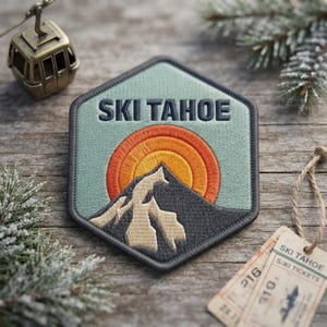 May include: A hexagonal embroidered patch featuring the text "SKI TAHOE" in dark blue letters above a dark gray mountain peak. Behind the mountain is a semi-circular sun design with concentric rings of orange and yellow thread on a sage green background.