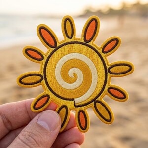 May include: An embroidered sun patch featuring a central spiral design in shades of yellow and cream. The sun has twelve radiating petals outlined in dark brown, with alternating orange and yellow centers, creating a circular sunburst motif.