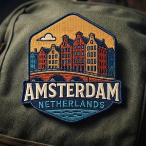 May include: A hexagonal embroidered patch featuring a colorful Amsterdam cityscape with canal houses, a bridge, and a single cloud against a yellow sky. The text AMSTERDAM is in large cream letters, with NETHERLANDS in smaller blue letters below.
