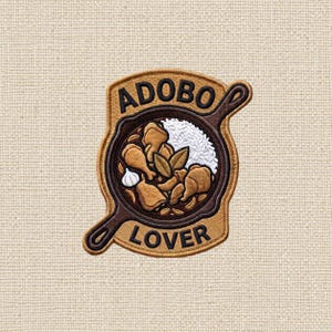 May include: An embroidered patch featuring a brown cast iron skillet filled with chicken pieces, white rice, garlic, and bay leaves. The text ADOBO is stitched in black above the skillet, and the word LOVER is stitched in black below the skillet.