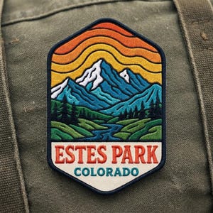 May include: A hexagonal embroidered patch featuring a landscape of blue mountains with white snow-capped peaks, a green forest, and a river. The sky is composed of orange and yellow wavy lines. The text ESTES PARK COLORADO is printed at the bottom.