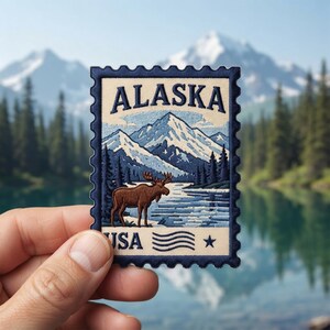 May include: A rectangular embroidered patch shaped like a postage stamp featuring a brown moose standing near a blue lake with snow-capped mountains and pine trees in the background. The text ALASKA is at the top and USA is at the bottom.