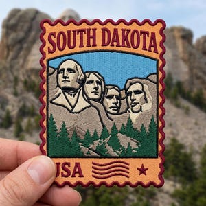 May include: A rectangular embroidered patch shaped like a postage stamp with a scalloped maroon border. It features the Mount Rushmore monument in tan and brown, green pine trees, a blue sky, and the text SOUTH DAKOTA at the top and USA at the bottom.