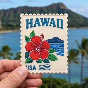 May include: A rectangular embroidered patch shaped like a postage stamp with a cream background. It features the blue text HAWAII at the top and USA at the bottom, a large red hibiscus flower with green leaves, and a blue mountain and ocean graphic.