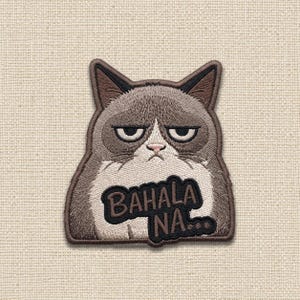 May include: An embroidered patch featuring a grumpy-looking cat with brown, gray, and white fur. The cat has a stern facial expression and is positioned above a black speech bubble containing the text BAHALA NA... in brown embroidered lettering.