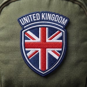May include: An embroidered patch featuring a shield shape with the Union Jack flag in red, white, and blue. The text UNITED KINGDOM is stitched in white capital letters on a dark blue arched banner at the top of the shield design.