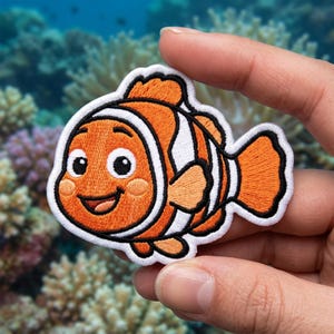 May include: An embroidered iron-on patch shaped like a clownfish with bright orange, white, and black thread. The fish has large eyes, a smiling mouth, and distinct white vertical stripes outlined in black, featuring a detailed stitched texture.