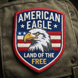 May include: A shield-shaped embroidered patch featuring a bald eagle head in profile with a yellow beak. The background displays the American flag with red and white stripes and blue with white stars. Text reads AMERICAN EAGLE LAND OF THE FREE.