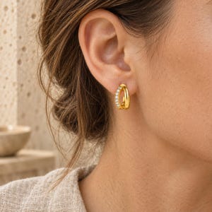 May include: A small gold hoop earring featuring a double-band design. One band is smooth and polished gold, while the other band is set with a row of small, clear, round-cut stones that sparkle against the metallic finish of the earring.