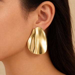 May include: A large, gold-toned earring with a smooth, polished surface and an irregular, organic, teardrop-like shape. The metal has a reflective finish with gentle curves that catch the light, creating subtle highlights across the surface.