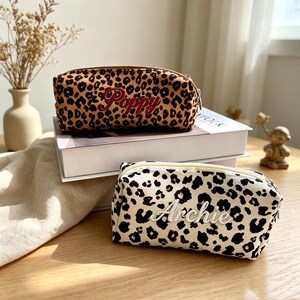 Könnte beinhalten: Zwei rechteckige Reißverschlusstaschen mit Leopardenmuster. Die obere Tasche ist braun und schwarz mit dem in Rot gestickten Namen Poppy. Die untere Tasche ist weiß und schwarz mit dem in weißer Schreibschrift gestickten Namen Archie.