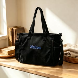 Puede incluir: Un bolso tote de pana negra con dos asas superiores y una correa de hombro desmontable. La parte frontal del bolso muestra el nombre Nelson bordado en hilo azul. Incluye un bolsillo lateral con cremallera vertical y compartimentos abiertos.