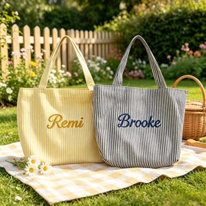 Puede incluir: Dos bolsas de tela con rayas verticales. La bolsa de la izquierda es amarilla con rayas blancas y tiene el nombre Remi bordado en hilo amarillo. La bolsa de la derecha es negra con rayas blancas y tiene el nombre Brooke bordado en hilo azul oscuro.