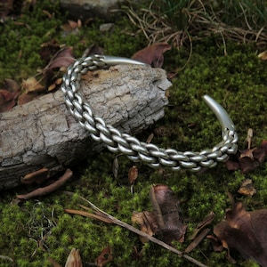 Könnte beinhalten: Ein silbernes Armband mit geflochtenem Design. Das Armband liegt auf einem Stück Rinde, das mit Moos bedeckt ist.