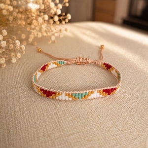 Puede incluir: Una pulsera tejida con cuentas que presenta un patrón geométrico en colores rojo, blanco, dorado y turquesa. La pulsera está confeccionada con hilo beige claro e incluye un cierre deslizante ajustable con dos extremos rematados en cuentas.
