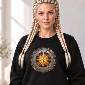 May include: A black long-sleeve sweatshirt featuring a circular graphic on the chest. The design displays a glowing orange and yellow lightning effect behind a silver rune symbol, encircled by a border of runic text. The text SIGNS & SYMBOLS is printed in small gold letters on the left sleeve.