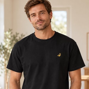 May include: A black short-sleeved t-shirt featuring a small, embroidered yellow scorpion graphic on the left chest. The garment has a classic crew neckline and a straight hem, constructed from a solid-colored fabric with a simple design.