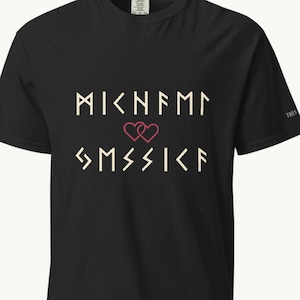 Pode incluir: Uma t-shirt preta de manga curta com duas linhas de texto em estilo rúnico na cor creme, separadas pelo contorno de um pequeno coração vermelho. O texto está centrado no peito e a t-shirt tem gola redonda e corte padrão.