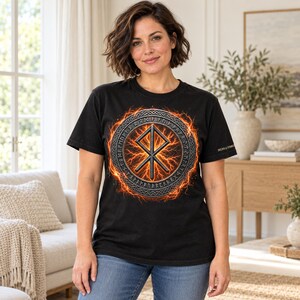 Pode incluir: Uma t-shirt preta com um gr&aacute;fico central de uma runa Othala met&aacute;lica dentro de uma borda circular de caracteres r&uacute;nicos, rodeada por um efeito de rel&acirc;mpagos laranja e amarelo. O texto SIGNS & SYMBOLS est&aacute; impresso na manga.
