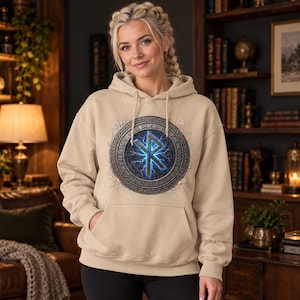 May include: A beige hooded sweatshirt featuring a central circular graphic with a glowing blue rune symbol surrounded by an intricate metallic border with etched characters. The hoodie includes a front kangaroo pocket and matching drawstrings.