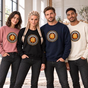 May include: A collection of four long-sleeved crewneck sweatshirts in dusty rose, black, navy blue, and cream. Each sweatshirt features a circular graphic on the chest displaying a glowing orange and yellow runic symbol with a dark border.