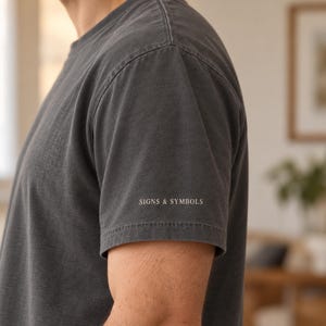 May include: A charcoal grey short-sleeved t-shirt with a crew neckline. The fabric has a slightly faded, washed appearance. The text "SIGNS & SYMBOLS" is printed in a small, clean, white sans-serif font on the lower edge of the left sleeve.