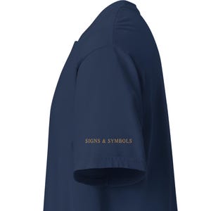 May include: A dark navy blue short-sleeved t-shirt viewed from the side. The sleeve features the text SIGNS & SYMBOLS printed in a metallic gold font. The garment has a simple, clean construction with a standard hemline on the sleeve and body.