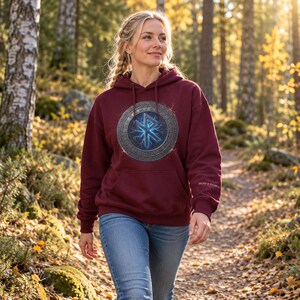 May include: A maroon hooded sweatshirt featuring a central circular graphic with a blue glowing Othala rune surrounded by a metallic-textured border. The hoodie includes a front kangaroo pocket and the text SIGNS & SYMBOLS on the left sleeve.