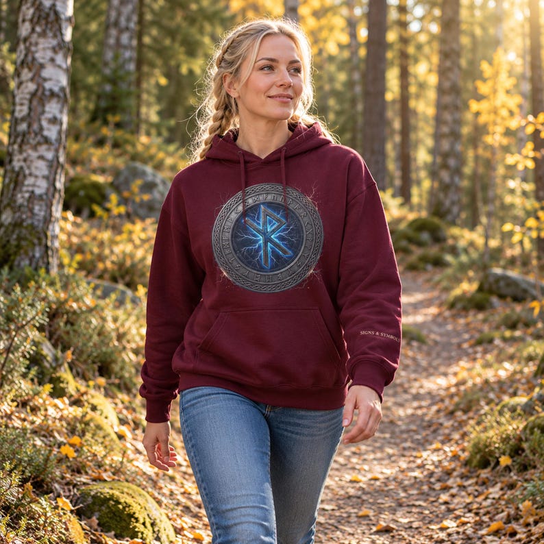 May include: A maroon hooded sweatshirt featuring a central circular graphic with a blue glowing Othala rune surrounded by a metallic-textured border. The hoodie includes a front kangaroo pocket and the text SIGNS & SYMBOLS on the left sleeve.