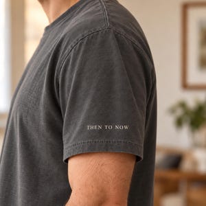 May include: A charcoal gray short-sleeve t-shirt featuring the text THEN TO NOW printed in a small, white, serif font on the lower portion of the left sleeve. The garment has a crew neckline and a visible seam along the shoulder and sleeve.