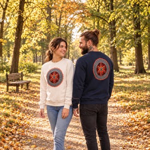 May include: A white crewneck sweatshirt and a navy blue crewneck sweatshirt, both featuring a circular graphic on the chest or back. The graphic displays a red and orange six-pointed star design surrounded by a detailed, intricate grey border.